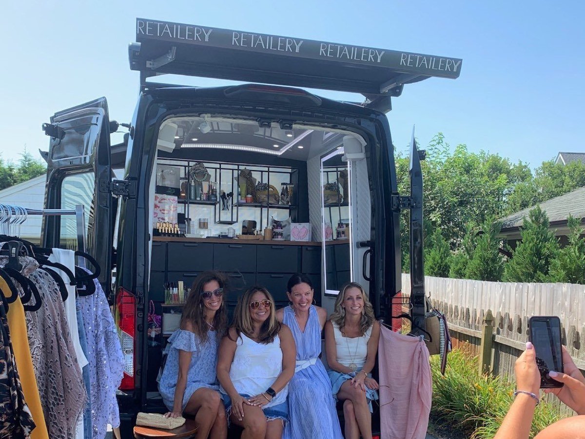 The Retailery mobile boutique van parked at an outdoor event, rear doors open with clothing racks and shelves of product, and four customers seated in the back.