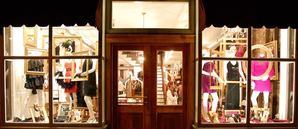 A boutique storefront lit up at night, its two display windows styled with mannequins in eveningwear and gilded picture frames, either side of a wooden double door.
